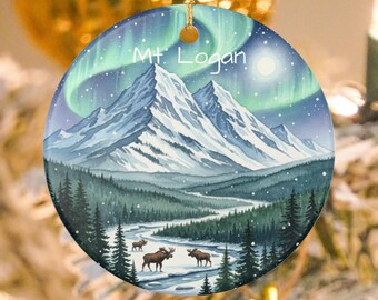 May include: Round Christmas ornament featuring a watercolor painting of a snowy mountain landscape with the text "Mt. Logan". The design includes a river, evergreen trees, moose, and an aurora borealis in the sky. The ornament is suspended by a gold string.