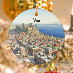 May include: Round ceramic ornament featuring a watercolor painting of a city with a large fortress on a hill overlooking a body of water. The word "Van" is printed at the top. The ornament has a gold string for hanging.