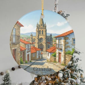 May include: Round Christmas ornament featuring a watercolor painting of a European town square with a cathedral. The buildings have red tile roofs and stone walls. The word "Guarda" is printed at the bottom.