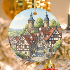 May include: Round ceramic ornament featuring a watercolor painting of Bad Wimpfen, Germany. The image depicts a quaint European town with half-timbered houses, red tile roofs, and two tall towers. The text "Bad Wimpfen" is visible at the bottom.