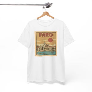May include: White t-shirt featuring a vintage-style graphic of Faro, Portugal. The design includes the word "FARO" above a red sun, with a detailed illustration of buildings, water, and boats in shades of beige, brown, and blue.
