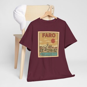 May include: Burgundy t-shirt featuring a vintage-style illustration of Faro, Portugal, with the city's name in a rectangular frame. The design includes a coastal scene with buildings, boats, and a setting sun. A white cap rests on a wooden chair.
