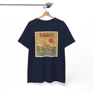 May include: Navy blue t-shirt featuring a vintage-style graphic of Faro, Portugal. The design includes the word "FARO" above a red sun, with a coastal town scene below. The shirt is hanging on a wooden hanger.