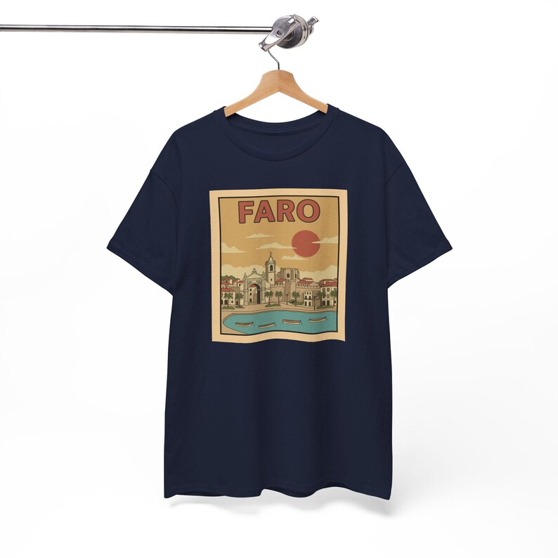 May include: Navy blue t-shirt featuring a vintage-style graphic of Faro, Portugal. The design includes the word "FARO" above a red sun, with a coastal town scene below. The shirt is hanging on a wooden hanger.