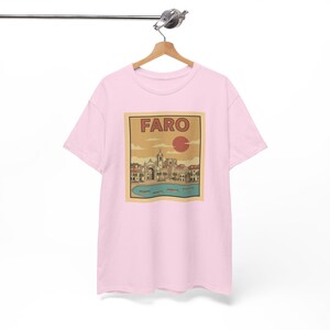 May include: A light pink t-shirt featuring a rectangular graphic with the word "FARO" in bold letters. The graphic depicts a coastal town scene with buildings, a church, and a red sun. The shirt is hanging on a wooden hanger.