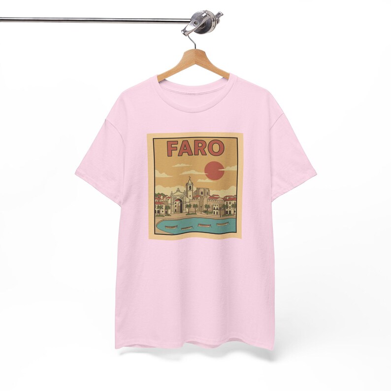 May include: A light pink t-shirt featuring a rectangular graphic with the word "FARO" in bold letters. The graphic depicts a coastal town scene with buildings, a church, and a red sun. The shirt is hanging on a wooden hanger.