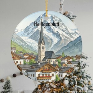 以下が含まれることがあります： オーストリアのハイリゲンブルットの水彩画が描かれた丸いクリスマスオーナメント。教会、村、雪をかぶった山々がデザインされています。「Heiligenblutt」の文字が上部に印刷されています。吊り下げ用の金色の紐が付いています。