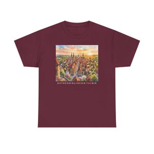 May include: A maroon t-shirt featuring a square watercolour illustration of the town of Rothenburg ob der Tauber at sunset. The graphic depicts historic buildings with red roofs, church spires, and trees, with the text ROTHENBURG OB DER TAUBER below.