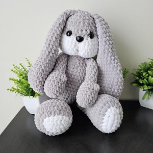 May include: A gray crocheted bunny with white accents on the ears and feet. The bunny is sitting on a dark gray surface.