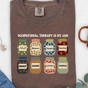 Puede incluir: Una camiseta de manga corta marrón con el texto "OCCUPATIONAL THERAPY IS MY JAM" sobre ocho frascos ilustrados. Cada frasco tiene un patrón diferente y una etiqueta con palabras como Adapt, Empower, Strength, Function, Engage, Achieve, Support y Progress.