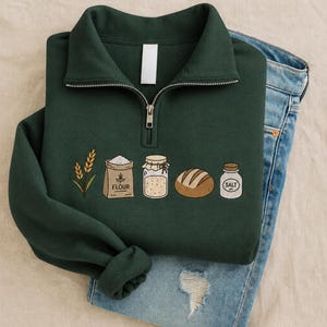 May include: A dark green quarter-zip sweatshirt featuring an embroidered graphic of wheat stalks, a brown bag labeled FLOUR, a glass jar, a loaf of bread, and a small white jar labeled SALT. The sweatshirt is layered over light blue denim jeans.