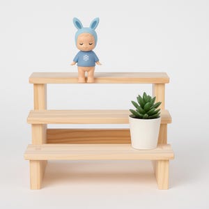 Könnte beinhalten: Ein dreistufiger Holzständer zeigt eine kleine blaue Figur mit Hasenohren und einem Schneeflockenmuster. Auf der untersten Ebene steht eine kleine Topf-Sukkulente. Der Ständer ist aus hellem Holz gefertigt.