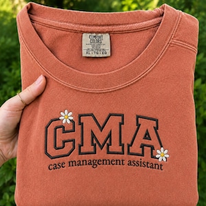 May include: A terracotta-colored crewneck sweatshirt featuring the embroidered black text CMA and case management assistant. The design includes two small white daisies with yellow centers. A Comfort Colors brand label is visible at the neckline.