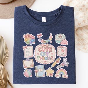 May include: A navy blue t-shirt with a collection of colorful, whimsical graphics. The design includes a pop-it toy, a beaded necklace, a container of putty, a clock, a pencil, scissors, a rainbow, and the text "CUTE OT THINGS".