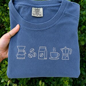 Puede incluir: Una sudadera azul de cuello redondo con un gráfico de líneas blancas que muestra una cafetera de goteo, tres granos de café, una bolsa de café, una taza de café humeante y una cafetera moka. La etiqueta interior indica Comfort Colors talla XL.