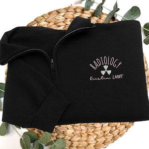 May include: Black quarter-zip sweatshirt featuring the word "RADIOLOGY" and a radiation symbol embroidered in light pink. The name "Kristin, LMRT" is embroidered below the symbol. The sweatshirt is displayed on a woven basket.