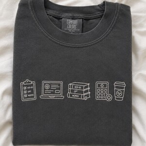Peut inclure: Un t-shirt à col rond gris anthracite avec un graphisme en lignes blanches représentant un presse-papiers avec des cases à cocher ICD-10, CPT et HCPCS, un ordinateur portable, trois livres étiquetés ICD-10, CPT et HCPCS, une calculatrice et un gobelet.