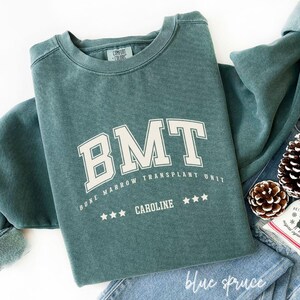 May include: A blue spruce sweatshirt with the text "BMT BONE MARROW TRANSPLANT UNIT CAROLINE" in white block letters. The sweatshirt is folded on top of blue jeans, with a pine cone and a small decorative bag.