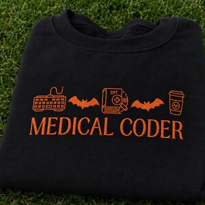 Puede incluir: Una sudadera negra de cuello redondo con bordado naranja. El diseño incluye un teclado, un murciélago, un libro con la etiqueta CPT y una lupa, otro murciélago y un vaso de café. El texto MEDICAL CODER está bordado en naranja debajo de los iconos.