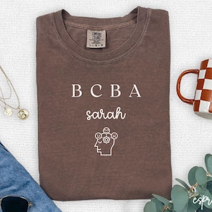 Peut inclure: Un t-shirt marron avec le texte "BCBA Sarah" et un graphique d'une tête avec des bulles de pensée. Les bulles de pensée ont différentes expressions faciales.