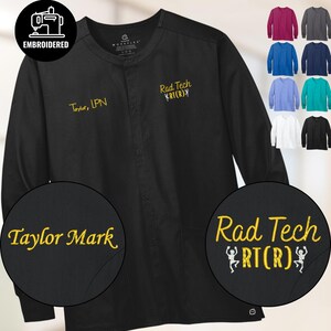 May include: Black long-sleeve medical scrub jacket with embroidered text. The jacket features the text "Taylor, LPN" and "Rad Tech RT(R)" in yellow thread. Additional embroidered text reads "Taylor Mark" and "Rad Tech RT(R)". A variety of colored scrub tops are displayed.