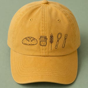 May include: A mustard yellow baseball cap featuring a black line art graphic on the front. The design consists of a loaf of bread, a small jar, a stalk of wheat, a whisk, and a spatula arranged in a horizontal row across the center of the cap.