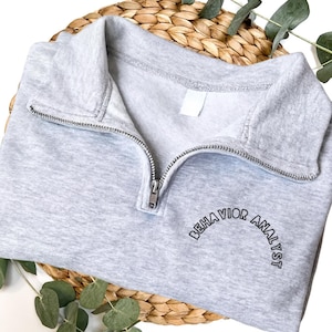 Puede incluir: Una sudadera gris claro con cremallera de cuarto de cremallera con el texto "BEHAVIOR ANALYST" en un diseño curvo. La sudadera tiene una cremallera plateada. La sudadera está doblada sobre una cesta tejida.