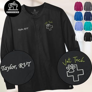 May include: Black long-sleeve medical scrub jacket with embroidered text "Taylor, RVT" and "Vet Tech" with a paw print and cross logo. Additional color options are displayed in the background. The jacket has a button-up front and a round neckline.