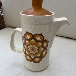 May include: A white ceramic coffee pot with a brown lid and a brown, orange, and white geometric design. The pot has a speckled glaze.