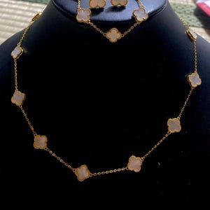 Op de afbeelding: Een goudkleurige set bestaande uit een ketting en bijpassende oorknopjes met klaverbladmotieven. Elk klaverblad is ingelegd met een glad, iriserend wit schelpmateriaal en omlijst door een verfijnde, kralenachtige goudkleurige metalen rand.