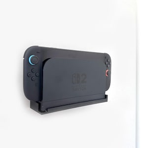 Op de afbeelding: Een zwarte Nintendo Switch-console en dock zijn op een witte muur gemonteerd. De console heeft blauwe en rode controllers. De dock heeft het Nintendo Switch-logo.