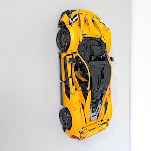 May include: A yellow and black model sports car made of interlocking plastic bricks. The car features black wheels, a black interior, and detailed exterior components. The model is mounted on a white wall, showcasing its design.