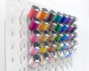 May include: A white pegboard with a variety of colored spools of thread organized in rows. The spools are arranged in a diagonal pattern, with the colors transitioning from light to dark.