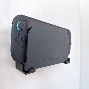 Op de afbeelding: Een zwarte gameconsole met een blauwe joystick en rode knoppen is aan een witte muur bevestigd. De console zit in een zwarte houder, ontworpen om het apparaat veilig en toegankelijk te houden. Het algehele ontwerp is strak en modern.