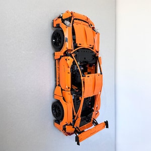 May include: An orange Lego model car mounted on a gray wall. The car has black wheels and headlights. The model is a detailed replica of a sports car.