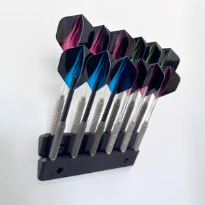 Może przedstawiać: Czarny uchwyt na rzutki z rzędem rzutek. Rzutki mają srebrne trzonki, czarne trzonki i kolorowe lotki w odcieniach niebieskiego, różowego i zielonego. Uchwyt jest zamontowany na białej ścianie.