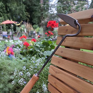 May include: A hand-held garden hoe featuring a triangular metal blade attached to a long, slender, dark metal neck. The tool has a smooth, light-colored wooden handle with a small hole at the base, through which a thin, light-colored cord is looped.