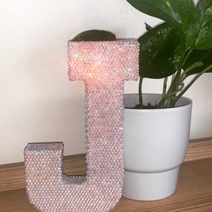 May include: A large, pink, glittery letter J made of rhinestones. The letter is sitting on a wooden surface next to a potted plant.