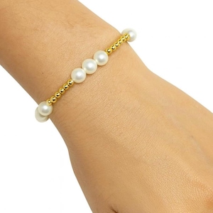 May include: A gold bracelet featuring a string of white pearls and small gold beads. The bracelet is worn on a wrist, showcasing the jewelry's design and color contrast. The pearls are evenly spaced, creating a classic and elegant look.