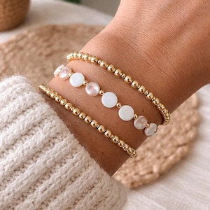 May include: Three gold beaded bracelets on a wrist. One bracelet features white, circular beads. The bracelets are stacked and worn over a cream-colored sweater. The background is a woven, tan surface.