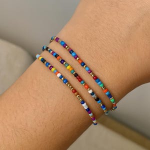 May include: Three colorful beaded bracelets with gold accents. The bracelets feature a variety of small, round beads in shades of blue, purple, yellow, orange, red, and white. The bracelets are strung together on a thin, flexible cord.