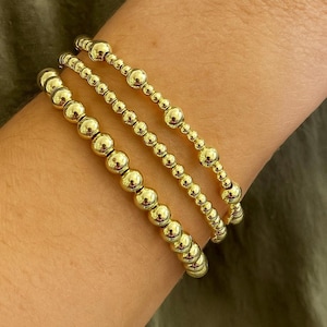 以下が含まれることがあります： 3つのゴールドビーズブレスレット。ブレスレットは、さまざまなサイズの丸く光沢のあるゴールドビーズでできています。ブレスレットは互いの上に積み重ねられており、レイヤードルックになっています。背景は無地のオリーブグリーンです。