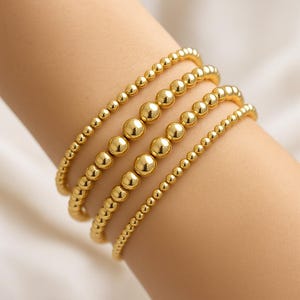 May include: Four gold beaded bracelets of varying sizes are stacked on a wrist. The bracelets feature small, round beads in a shiny gold color. The bracelets are a simple, elegant accessory.