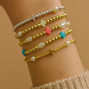 May include: A collection of beaded bracelets in gold and silver tones. The bracelets feature small beads, crosses, and pearls in various colors. The bracelets are stacked on a wrist, showcasing the different designs and textures.