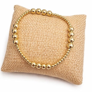 May include: A gold beaded bracelet with varying sized beads. The bracelet is displayed on a small, square, tan burlap pillow. The beads are round and shiny, reflecting light. The bracelet is a simple, elegant piece of jewelry.