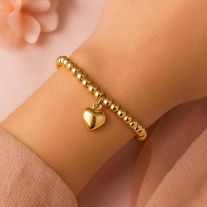Op de afbeelding: Gouden kralenarmband met een hartvormige bedel. De armband heeft kleine, ronde kralen en een gepolijste hartvormige hanger. De sieraden worden op een pols getoond, waarbij het ontwerp en de kleur worden getoond.