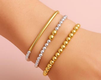 May include: Three bracelets on a wrist. One is a gold bar, the other two are beaded, one silver and gold, the other all gold. The background is a solid light pink.