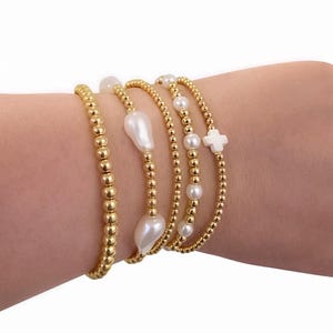 May include: Gold beaded bracelets with freshwater pearls and a white cross charm. The bracelets are worn on a wrist and are made of small, round gold beads. Some bracelets have large, irregular-shaped pearls.