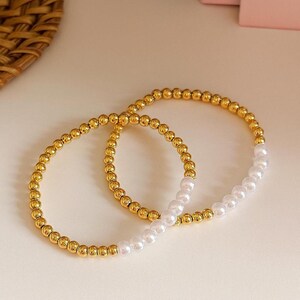 May include: Two gold beaded bracelets with a section of white pearl beads. The bracelets are circular and sit on a light-colored surface. The gold beads have a shiny finish, and the pearls are smooth and round. The bracelets are a jewelry item.