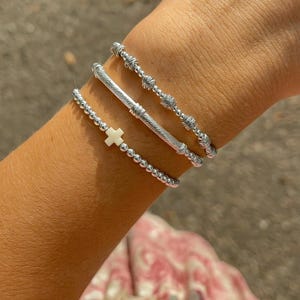 Peut inclure: Plusieurs bracelets argentés. L'un est orné d'une croix blanche, un autre d'une barre texturée, et les autres de perles. Les bracelets sont portés sur un poignet.
